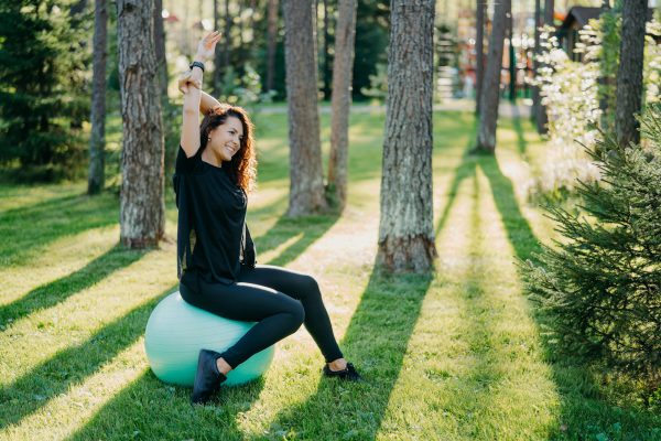 Relaxed brunette woman sits on fitness ball raises arms and does excercises outdoor